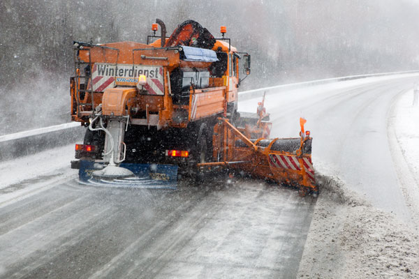 Ein orangefarbener Schneepflug mit der Aufschrift 'Winterdienst' räumt den Schnee von einer verschneiten Autobahn. Das Fahrzeug sprüht Salz und hat rot-weiße Warnmarkierungen. Der Himmel ist bewölkt und es schneit leicht. Der Einsatz dient der Verkehrssicherheit und gehört zur präventiven Straßenwartung im Winterdienst.