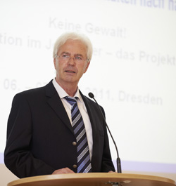 Ein älterer Mann mit weißem Haar und Brille steht an einem Rednerpult und hält eine Rede. Er trägt einen dunklen Anzug mit gestreifter Krawatte. Im Hintergrund ist eine Projektionstafel zu sehen, auf der unscharf Text zur Gewaltprävention erkennbar ist. Der Kontext bezieht sich auf Arbeitsschutz und Präventionsmaßnahmen, relevant für die Unfallkasse NRW.