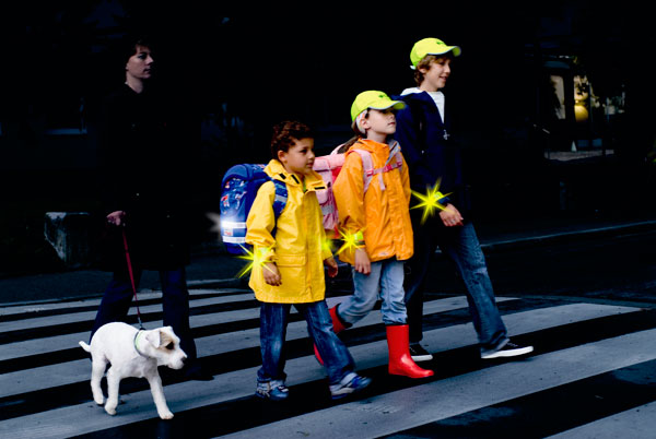 Zwei Erwachsene und zwei Kinder mit Hund überqueren bei Dunkelheit einen Zebrastreifen. Die Kinder tragen reflektierende Kleidung, Mützen und Rucksäcke, die im Licht leuchten, um die Sichtbarkeit zu erhöhen. Das Bild thematisiert Arbeitsschutz und Prävention durch die Verwendung von Sicherheitskleidung im Verkehr, relevant für die Unfallkasse NRW.