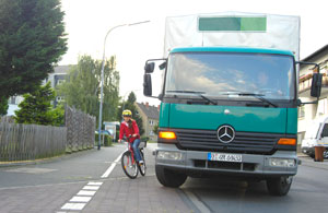 Ein grüner LKW überholt einen Radfahrer auf einer schmalen Straße in einem Wohngebiet. Der Radfahrer trägt Helm und Schutzkleidung. Die Szene betont die Wichtigkeit von Achtsamkeit und Vorsicht im Straßenverkehr, speziell in Bezug auf Arbeitsschutz und Prävention gemäß den Richtlinien der Unfallkasse NRW.
