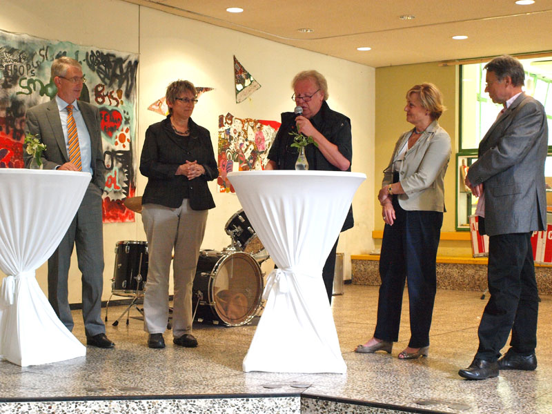 Fünf Personen stehen in einem Veranstaltungsraum mit gemusterten Wänden und einem Schlagzeug im Hintergrund. Zwei Stehtische mit weißen Tischdecken sind im Vordergrund platziert, auf denen kleine Vasen mit Blumen stehen. Eine Person spricht in ein Mikrofon und wird von den anderen aufmerksam betrachtet. Das Umfeld und die Personen deuten auf ein Event zur Arbeitssicherheit oder Prävention hin, im Kontext der Unfallkasse NRW.