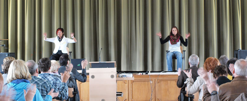 Zwei Personen, eine in weißem Blazer, die andere in gestreiftem Pullover, sitzen auf einer Bühne vor einem Publikum. Sie führen mit erhobenen Armen eine Übung vor. Die Bühne hat einen Tisch mit Mikrofon und Kabeln. Zuschauer klatschen und schauen auf die Bühne. Der Raum hat einen Vorhang im Hintergrund, der den Bühnenbereich abgrenzt. Die Szene könnte Teil eines Seminars oder Workshops zur Prävention und Sicherheit am Arbeitsplatz sein, organisiert von der Unfallkasse NRW.