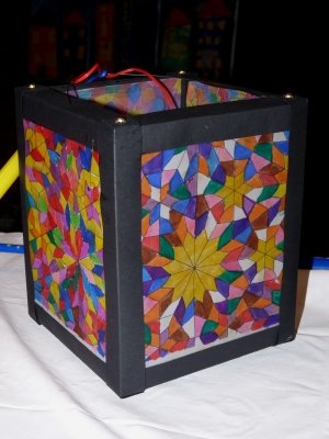 Eine quadratische Laterne aus schwarzem Karton enthält bunte, geometrische Glasfenstermuster auf den Seiten. Die Muster bestehen aus farbenfrohen, ineinander verschlungenen Formen, die an ein Kaleidoskop erinnern. Oben auf der Laterne sind Kabel sichtbar. Die Laterne steht auf einem weißen Untergrund, vor einem dunklen Hintergrund. Diese Art von Laterne kann in Kreativ-Workshops zur Förderung von Teamarbeit und Achtsamkeit im Kontext von Arbeitsschutz und Prävention verwendet werden.