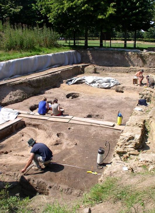 Mehrere Personen arbeiten in einer archäologischen Ausgrabungsstätte im Freien. Sie tragen Arbeitskleidung und sind mit Werkzeugen wie Kellen und Besen beschäftigt. Auf dem Boden sind verschiedene Ausgrabungsbereiche sichtbar. Die Szenerie ist von grünem Bewuchs umgeben, und im Hintergrund sind Bäume zu sehen. Die Ausgrabung ist durch Sicherheitsvorkehrungen wie Schutzzäune abgesichert, um Unfälle zu vermeiden.