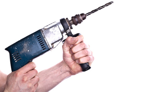 Nahaufnahme einer Hand, die eine elektrische Bohrmaschine festhält. Der Bohrer ist schwarz und die Maschine blau, mit einem abgenutzten Griff. Der Hintergrund ist weiß und schlicht gehalten, um den Fokus auf die sichere Handhabung des Werkzeugs zu legen. Konzipiert für den Einsatz im Kontext von Arbeitsschutz und Prävention durch die Unfallkasse NRW.