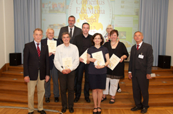 Acht Personen posieren für ein Foto auf einer Bühne. Einige halten Urkunden in den Händen, möglicherweise im Rahmen einer Veranstaltung zur Förderung von Arbeitsschutz und Prävention, organisiert von der Unfallkasse NRW. Im Hintergrund ist eine Projektion mit der Aufschrift 'Compensus FaMiLia' zu sehen.