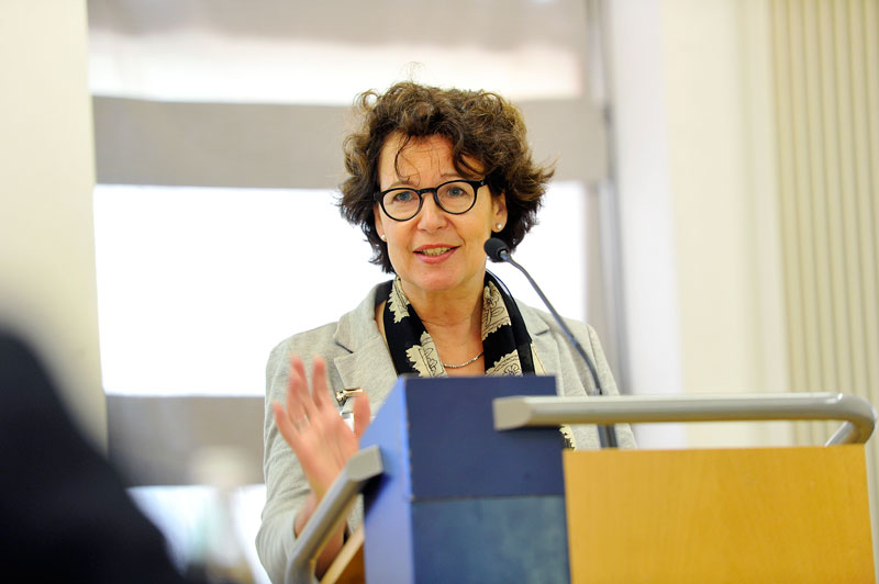 Eine Frau steht hinter einem Rednerpult und hält eine Präsentation. Sie trägt eine Brille und berufliche Kleidung. Im Hintergrund ist ein Fenster mit einem halb geöffneten Rollo zu sehen. Das Bild vermittelt einen formalen Kontext, der auf Arbeitsschutz und Prävention im Rahmen einer Veranstaltung der Unfallkasse NRW hindeutet.