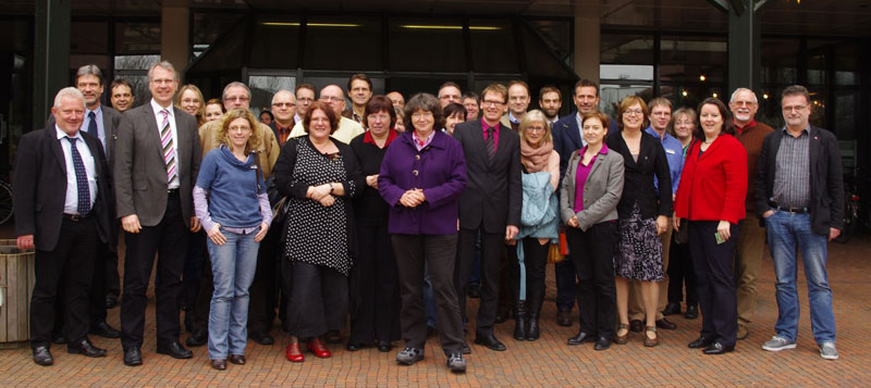 Eine diverse Gruppe von etwa 30 Personen steht vor dem Eingangsbereich eines modernen Gebäudes. Die Gruppe ist gemischt geschlechtlich und von unterschiedlichem Alter, in business-casual Kleidung. Einige Personen lächeln und stehen nah beieinander, was auf einen freundlichen Anlass hindeutet, möglicherweise ein Seminar oder eine Veranstaltung zur Arbeitssicherheit. Das Gebäude im Hintergrund ist modern mit Glas- und Metallelementen, im Innenbereich sind Fahrräder zu erkennen. Der Anlass ist höchstwahrscheinlich im Kontext von Arbeitsschutz und Prävention bei der Unfallkasse NRW.