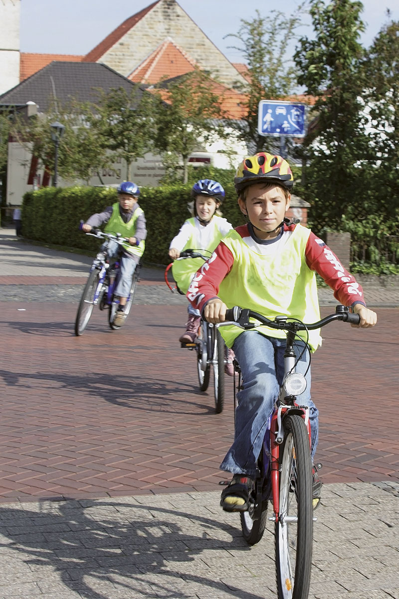 Drei Kinder fahren auf einem gepflasterten Weg Fahrrad. Sie tragen gelbe Sicherheitswesten und Helme. Der Vordergrund zeigt ein Kind mit rotem Fahrrad und rotem Helm, gefolgt von zwei weiteren Kindern. Im Hintergrund sind Gebäude mit roten Ziegeldächern und ein Verkehrszeichen zu sehen. Der Fokus liegt auf Sicherheit und Prävention im Straßenverkehr für Schulkinder, passend zum Kontext der Unfallkasse NRW.