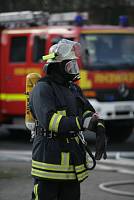 Ein Feuerwehrmann in voller Schutzkleidung mit Atemschutzmaske und Sauerstoffflasche steht auf einem Parkplatz vor einem roten Feuerwehrauto mit der Aufschrift 'Feuerwehr'. Er trägt Handschuhe und blickt zur Seite. Im Hintergrund sind weitere Ausrüstungsgegenstände und Fahrzeuge sichtbar. Das Bild betont die Wichtigkeit von Arbeitsschutz und Sicherheitsmaßnahmen im Einsatz, im Kontext von Präventionsmaßnahmen der Unfallkasse NRW.