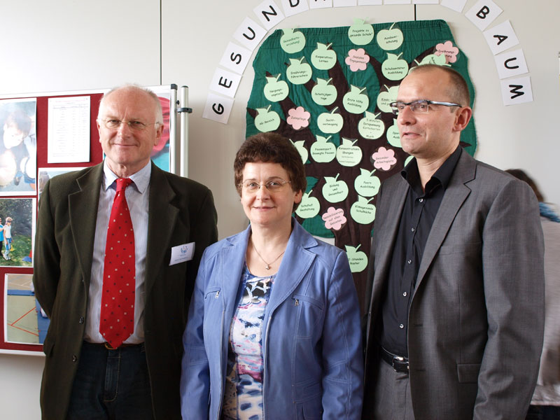 Drei Personen posieren nebeneinander vor einem Plakat mit der Überschrift 'Gesundheitsbaum'. Auf dem Plakat sind verschiedene Blätter mit Gesundheitsbegriffen befestigt. Die Personen tragen formelle Kleidung und stehen in einem Raum, der Arbeits- und Gesundheitsschutz thematisiert. Das Bild vermittelt den Kontext eines Gesundheits- oder Präventionsprojekts bei der Unfallkasse NRW.