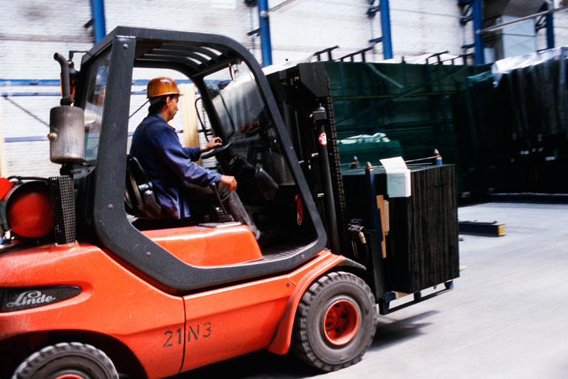 Ein Mann mit Sicherheitshelm fährt einen roten Gabelstapler und transportiert vorsichtig mehrere große Glasplatten in einer geräumigen Lagerhalle. Die Glasplatten sind sicher auf der Gabel des Fahrzeugs befestigt. Die Szene verdeutlicht die Wichtigkeit von Arbeitsschutz und ordnungsgemäßer Ladungssicherung im Lagerbetrieb.