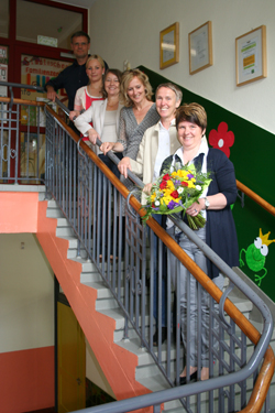 Sechs Personen stehen lächelnd hintereinander auf einer Treppe in einem Gebäude. Die Person vorne hält einen bunten Blumenstrauß. Die Treppe ist mit einem Metallgeländer gesichert und die Umgebung ist mit einem grünen Wandgemälde und Rahmen dekoriert. Dies könnte im Kontext der Unfallkasse NRW für Arbeitsschutz und Prävention stehen, da es auf gute Zusammenarbeit und Anerkennung hindeutet.
