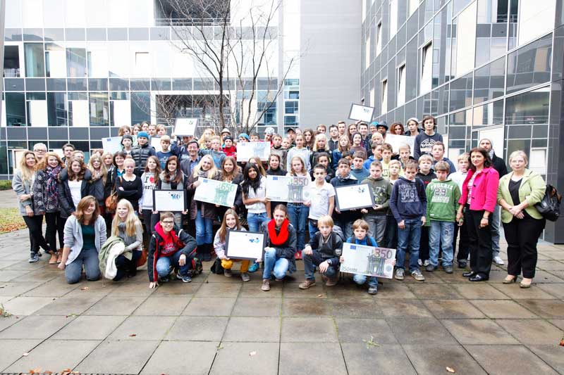 Eine große Gruppe von Schülern in Freizeitkleidung hält Zertifikate in den Händen, während sie vor einem modernen Bürogebäude posiert. Der Hintergrund zeigt eine Glasfassade und kahle Bäume. Erwachsene, vermutlich Lehrkräfte oder Mitarbeiter der Unfallkasse NRW, stehen am Rand der Gruppe und lächeln. Die Szene steht im Kontext der Arbeitsschutz- und Präventionsmaßnahmen, die die Unfallkasse NRW unterstützt.