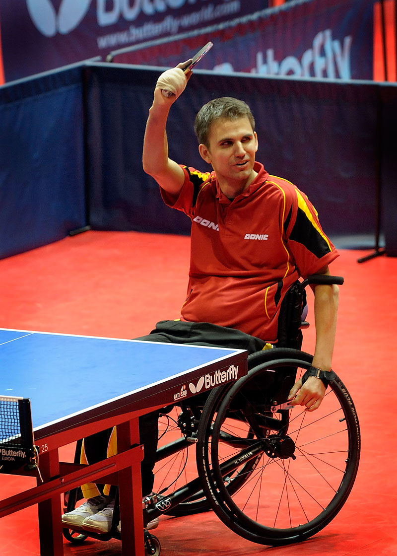 Ein männlicher Rollstuhlfahrer spielt Tischtennis in einer Sporthalle. Er trägt ein rotes Sporttrikot und hebt triumphierend seinen Tischtennisschläger in die Luft. Die Szene vermittelt eine positive Stimmung des Sieges und der Freude am Sport. Der Hintergrund zeigt einen Tischtennistisch und eine blaue Spielfeldumrandung. Kontext: Förderung von Inklusion und Prävention durch die Unfallkasse NRW.