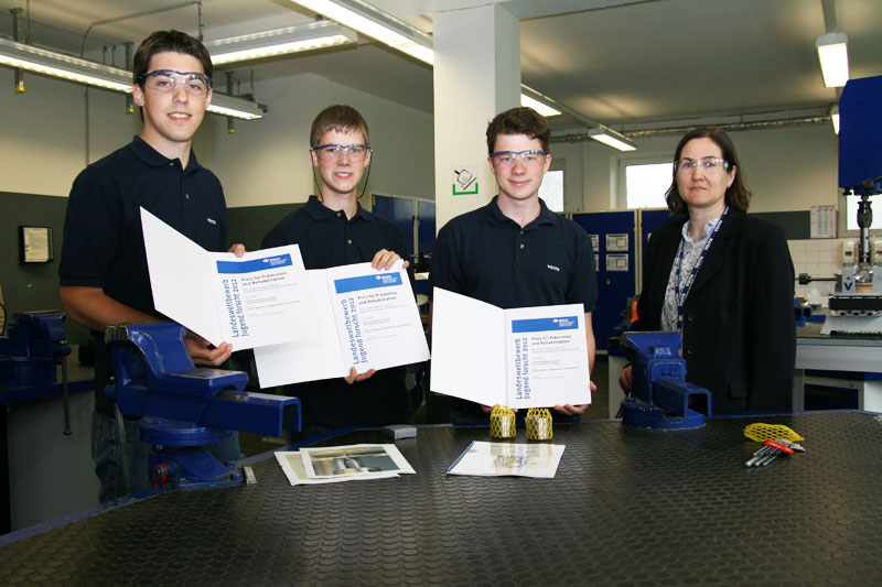 Drei junge Männer in blauen Poloshirts und Schutzbrillen stehen in einer Werkstatt und halten Urkunden der Unfallkasse NRW. Neben ihnen steht eine Frau in einem Blazer. Auf dem Tisch liegen Werkzeuge und gelbe Arbeitshandschuhe. Der Raum ist gut beleuchtet mit Maschinen im Hintergrund. Kontext ist die Förderung von Sicherheit und Prävention am Arbeitsplatz.