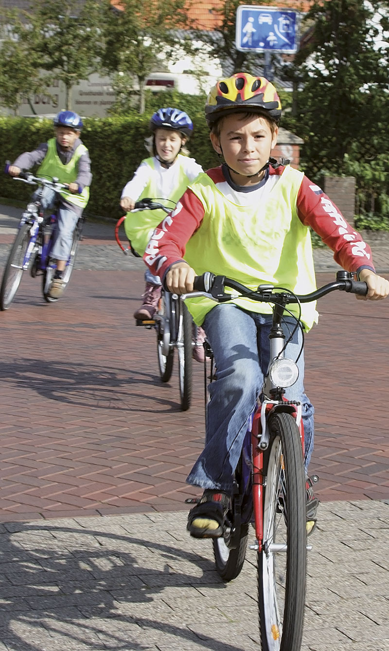 Drei Kinder fahren mit Fahrrädern auf einem gepflasterten Weg. Sie tragen gelbe Warnwesten und Helme als Sicherheitsmaßnahme. Die Umgebung zeigt Bäume und Gebäude im Hintergrund. Das Bild vermittelt eine Botschaft der Sicherheit und Prävention im Straßenverkehr, passend zum Thema Arbeitsschutz und Unfallprävention der Unfallkasse NRW.