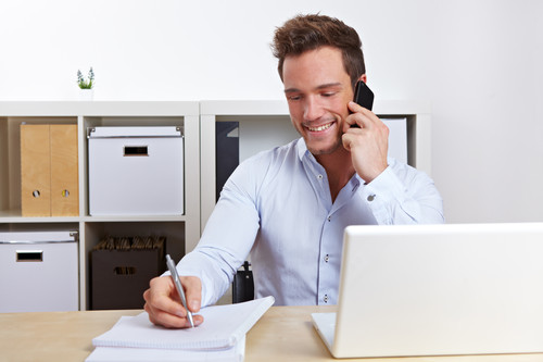 Ein Mann in einem weißen Hemd sitzt an einem Schreibtisch in einem Büro. Er hält ein Smartphone ans Ohr und macht Notizen auf einem Block. Vor ihm steht ein Laptop. Im Hintergrund sind weiße Regale mit Ordnern und Boxen zu sehen. Das Bild thematisiert Arbeitsschutz und effiziente Kommunikation in Büroumgebungen, passend zum Kontext der Unfallkasse NRW.