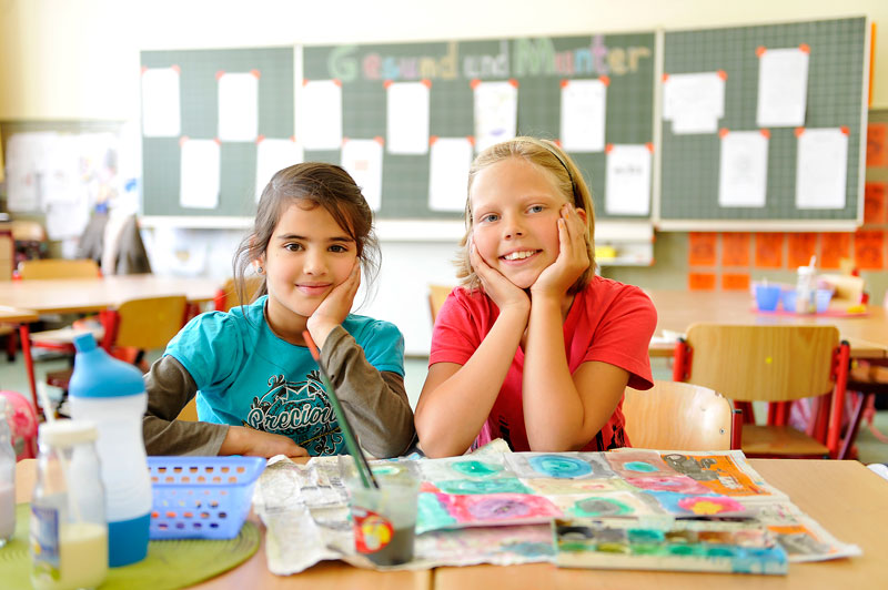 Zwei lächelnde Kinder sitzen an einem Tisch in einem Klassenzimmer und beschäftigen sich mit Malutensilien. Auf dem Tisch liegen Wasserfarben und Pinsel bereit, während im Hintergrund eine Pinnwand mit Dokumenten sichtbar ist. Die Szene vermittelt eine freundliche Lernumgebung und steht im Kontext von kreativem Lernen und Sicherheit im Unterrichtsalltag, wie es von der Unfallkasse NRW gefördert wird.