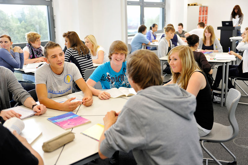 Eine Gruppe von Schülern sitzt an Tischen in einem modernen Klassenraum. Sie scheinen eine lebhafte Diskussion zu führen und sind in Kleingruppen aufgeteilt. Bücher und Schreibmaterialien liegen auf den Tischen. Im Hintergrund sind weitere Schüler zu sehen, die ebenfalls in Gruppen zusammenarbeiten. Durch die Fenster fällt Tageslicht in den Raum. Das Bild vermittelt einen Eindruck von aktivem, kooperativem Lernen. Es könnte in den Kontext von Arbeitsschutz und Prävention gestellt werden, da effektive Kommunikation und Gruppenarbeit Sicherheitsrisiken am Arbeitsplatz reduzieren können.