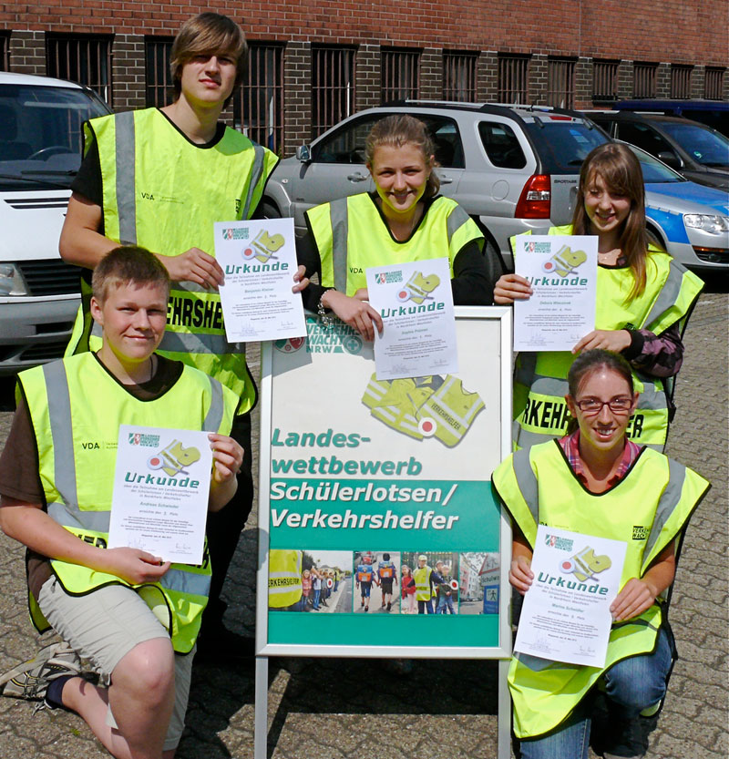 Fünf Jugendliche stehen auf einem Parkplatz und tragen gelbe Warnwesten. Sie halten jeweils eine Urkunde in der Hand. Vor ihnen steht ein Schild mit der Aufschrift 'Landeswettbewerb Schülerlotsen/Verkehrshelfer'. Im Hintergrund sind geparkte Autos und ein Backsteingebäude zu sehen. Die Szene betont die Bedeutung von Arbeitsschutz und Prävention im Straßenverkehr, im Kontext der Unfallkasse NRW.
