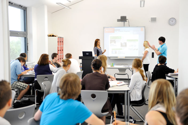 Ein Schulungsraum mit etwa 15 Personen, hauptsächlich junge Erwachsene, die an Tischen sitzen. Zwei junge Erwachsene stehen vorne und präsentieren Informationen auf einer Projektionsfläche. Eine Frau steht neben einem Rednerpult, vermutlich die Lehrkraft. Die Teilnehmer scheinen Notizen zu machen oder den Vortrag aufmerksam zu verfolgen. Das Szenario wirkt interaktiv und gut strukturiert, passend zum Kontext der Unfallkasse NRW, die Arbeitsschutz und Prävention thematisiert.