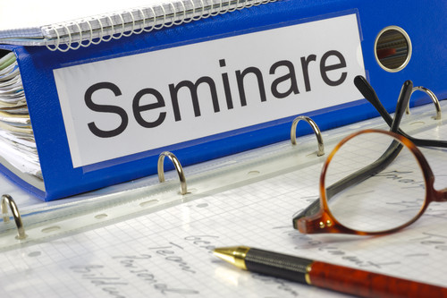 Ein blauer Ordner mit der Aufschrift 'Seminare' liegt auf einer Schreibunterlage mit kariertem Papier. Daneben befindet sich eine rote Brille und ein Kugelschreiber. Der Kontext deutet auf Bildungs- und Präventionsmaßnahmen der Unfallkasse NRW hin, die Arbeitsschutz und Sicherheit am Arbeitsplatz fördern sollen.