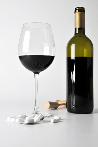 Ein Glas Rotwein steht gefüllt neben einer geöffneten Weinflasche und einem Blister mit weißen Tabletten auf einem hellen Untergrund. Diese Anordnung könnte auf potenzielle Gefahren und Risiken hinweisen, die durch die gleichzeitige Einnahme von Alkohol und Medikamenten entstehen können, was im Kontext von Arbeitsschutz und Prävention relevante Themen sind.