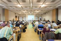 Großer, gut beleuchteter Seminarraum mit vielen Sitzplätzen, die von Teilnehmern besetzt sind. Alle Blicke sind auf eine Leinwand vorne gerichtet, auf der eine Präsentation angezeigt wird. Im Raum befinden sich Menschen verschiedenen Alters, die konzentriert der Präsentation folgen. Es wird ein Vortrag über Arbeitsschutz und Prävention gehalten, organisiert von der Unfallkasse NRW. Im Vordergrund stehen Mikrofone, die dafür sorgen, dass der Vortrag gut verständlich übertragen wird.