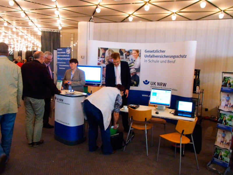 Der Infostand der Unfallkasse NRW ist auf einer Messe präsent. Mehrere Personen stehen an einem Tisch mit Computern und Informationsmaterial. Im Hintergrund sind Banner mit dem Logo der Unfallkasse NRW und dem Text 'Gesetzlicher Unfallversicherungsschutz in Schule und Beruf' sichtbar. Der Stand informiert über Arbeitsschutz und Prävention. Der Raum ist gut beleuchtet und es sind weitere Stände erkennbar.
