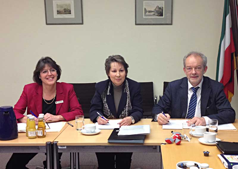 Drei Personen, zwei Frauen und ein Mann, sitzen an einem Holztisch in einem Konferenzraum. Auf dem Tisch befinden sich Dokumente, Schreibutensilien, eine blaue Kaffeekanne, Saftflaschen, Kaffeetassen und ein Plüsch-Maskottchen der Unfallkasse NRW. An der Wand hängen zwei Bilder und eine deutsche Flagge. Diese Szene könnte ein Treffen zur Besprechung von Arbeitsschutzmaßnahmen und Präventionsstrategien darstellen.