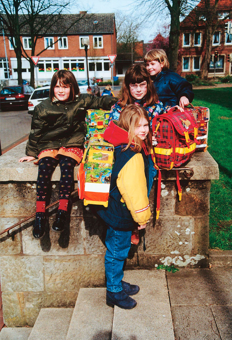 Vier Kinder mit bunten Schulranzen sitzen auf einer niedrigen Mauer am Straßenrand. Im Hintergrund sind geparkte Autos und Wohnhäuser zu sehen. Die Szene betont die Wichtigkeit von Arbeitsschutz und Prävention im Straßenverkehr, insbesondere für junge Schulkinder. Die Kinder sind warm gekleidet und scheinen gemeinsam ihren Schulweg zu beginnen.