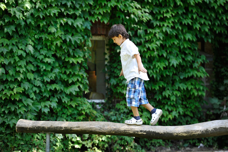 Ein junger Junge balanciert konzentriert auf einem waagerecht liegenden Baumstamm. Er trägt ein weißes Hemd, karierte Shorts und weiße Schuhe. Im Hintergrund ist eine mit Efeu bewachsene Wand zu sehen. Diese Szene symbolisiert Themen wie Geschicklichkeit, Gleichgewicht und möglicherweise Unfallprävention in der Natur. Der Kontext könnte auf sichere Spielumgebungen und Präventionsmaßnahmen im Sinne der Arbeitsschutzrichtlinien der Unfallkasse NRW hinweisen.