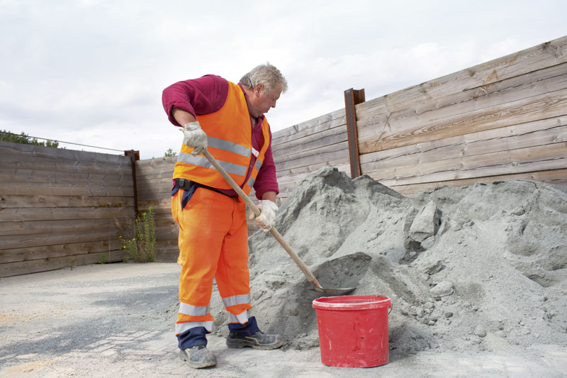 Ein älterer männlicher Bauarbeiter in orangefarbener Schutzkleidung mit reflektierenden Streifen schaufelt Sand aus einem großen Haufen in einen roten Eimer. Er trägt Handschuhe und steht in einem von Holzwänden umgebenen Bereich auf einer Baustelle. Diese Szene betont den Arbeitsschutz und die Prävention, da der Arbeiter angemessene Schutzkleidung trägt.