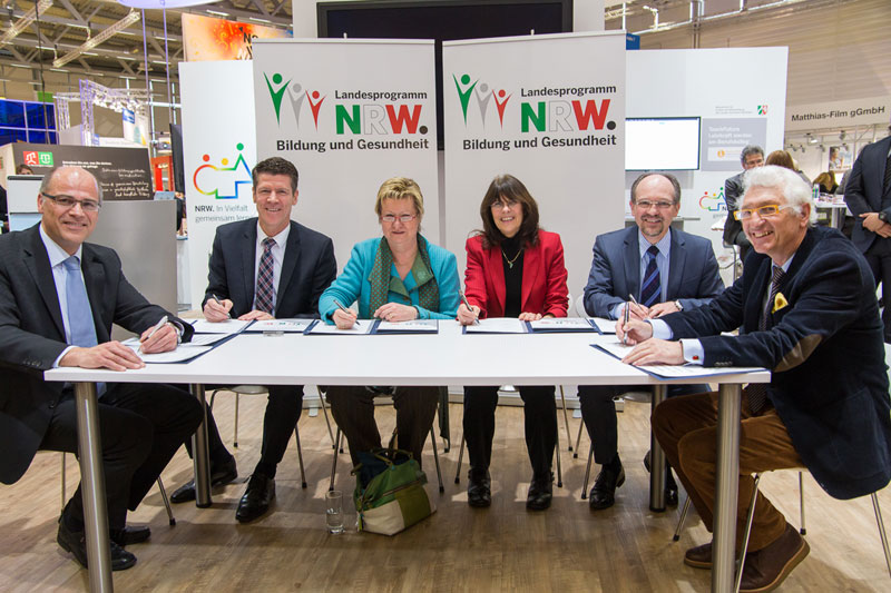 Sechs Personen sitzen an einem Tisch und unterzeichnen Dokumente. Im Hintergrund sind Logos des Landesprogramms NRW für Bildung und Gesundheit zu sehen. Die Szene deutet auf eine Zusammenarbeit hin, die sich vermutlich mit dem Thema Arbeitsschutz und Prävention befasst. Die Personen wirken offiziell und konzentriert. Die Umgebung ist eine Messe oder ein ähnlicher öffentlicher Raum mit Ständen und Informationsmaterialien.