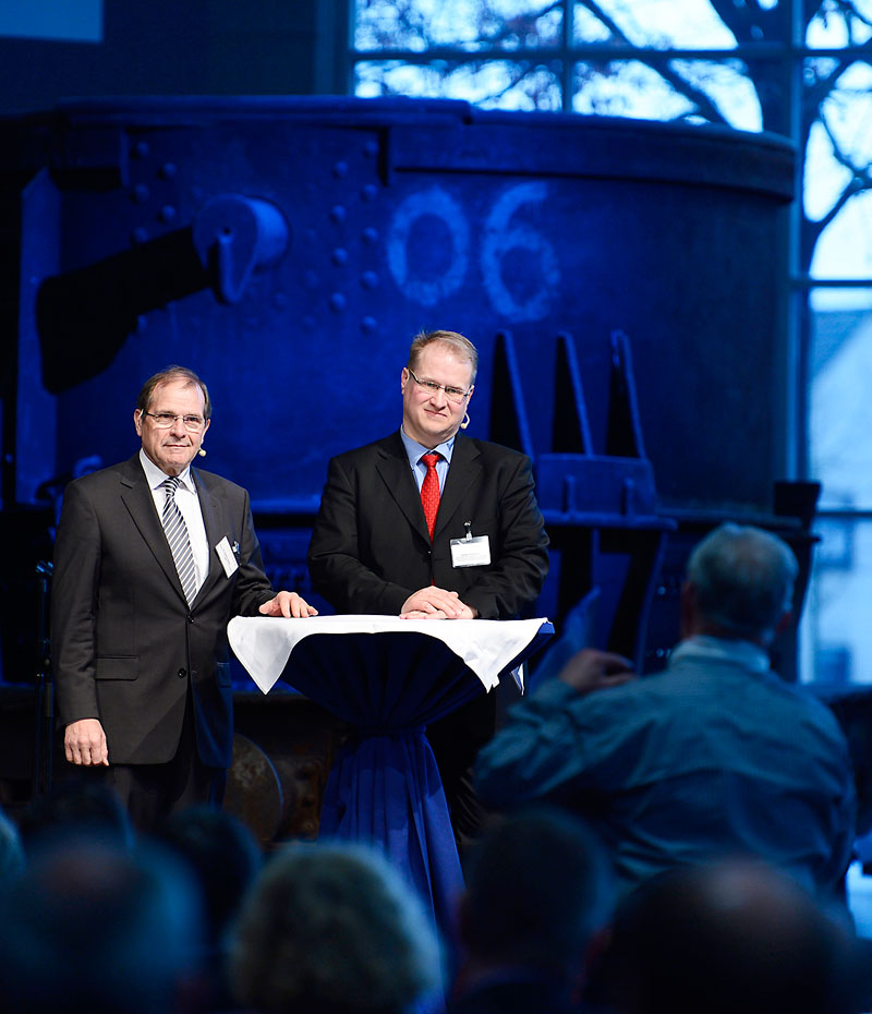 Zwei Männer in Anzügen stehen an einem mit einem weißen Tischtuch bedeckten Stehtisch. Einer trägt eine rot-schwarze Krawatte. Im Hintergrund ist eine große industrielle Anlage zu sehen, die beleuchtet ist. Vor ihnen sitzt ein Publikum, das den Vortrag oder die Diskussion verfolgt. Das Bild vermittelt ein professionelles Umfeld, das sich möglicherweise auf Arbeitsschutz oder Prävention bezieht, passend zum Kontext der Unfallkasse NRW.