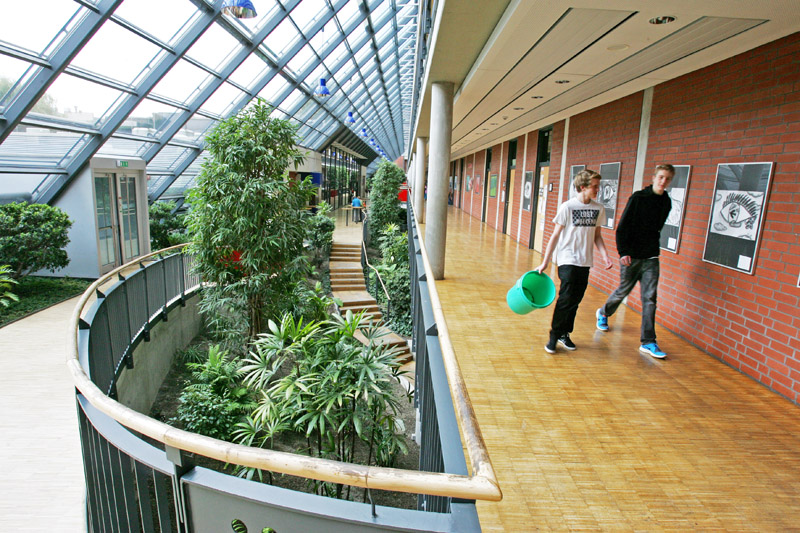Ein moderner Flur mit breitem Fensterdach und roten Ziegelwänden. Links ein Atrium mit üppigen Pflanzen. Rechts zwei Jugendliche in Freizeitkleidung, die jeweils einen grünen Eimer tragen. Kontext der Unfallkasse NRW: Der Fokus liegt auf der Sicherheit und Prävention im Arbeitsumfeld, hier beispielhaft in einer Bildungseinrichtung.