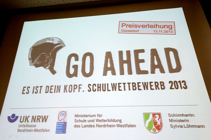 Ein Plakat des Schulwettbewerbs 2013, Go Ahead, thematisiert Arbeitsschutz und Prävention. Oben rechts steht 'Preisverleihung Düsseldorf 13.11.2013'. Zentral ein gezeichneter Schutzhelm mit dem Slogan 'GO AHEAD Es ist dein Kopf'. Unten sind Logos der Unfallkasse NRW und des Ministeriums für Schule und Weiterbildung des Landes Nordrhein-Westfalen zu sehen. Die Schirmherrin ist Ministerin Sylvia Löhrmann.