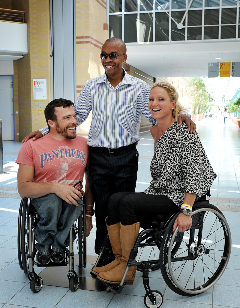 Das Bild zeigt drei lächelnde Personen in einem modernen, hellen Gebäude mit einer Glasfassade im Hintergrund. Zwei Personen sitzen in Rollstühlen, eine Person steht zwischen ihnen. Der Ort wirkt einladend und barrierefrei, was auf das Thema Inklusion und Barrierefreiheit im Kontext des Arbeitsschutzes und der Prävention bei der Unfallkasse NRW hinweist.