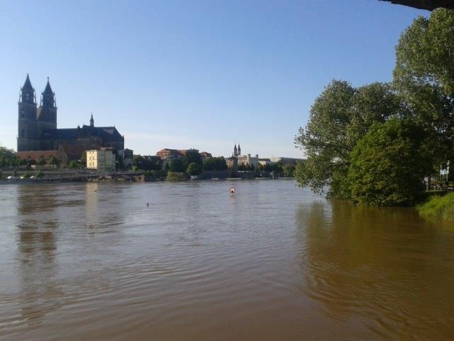 Das Bild zeigt einen breiten Fluss mit ruhigem Wasser und einer großen Kathedrale am gegenüberliegenden Ufer. Am rechten Ufer befinden sich Bäume. Das Wasser wirkt hoch, möglicherweise nach starken Regenfällen. Der Himmel ist klar und blau. Im Kontext der Unfallkasse NRW könnte dies auf Hochwasserschutz und Präventionsmaßnahmen hinweisen, um die Sicherheit der Umgebung zu gewährleisten.