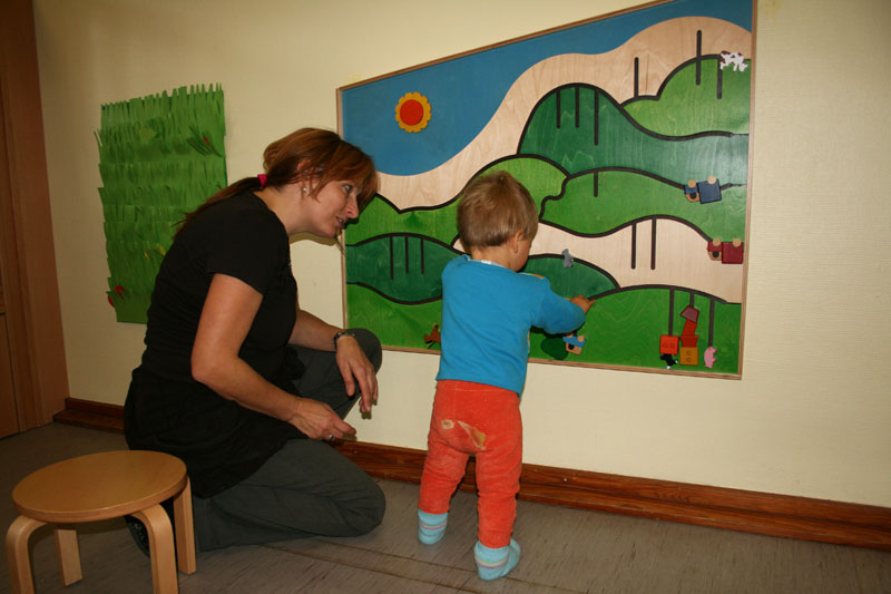 Eine Frau kniet neben einem Kleinkind, das an einem Wandspielbrett mit einem landschaftlichen Design spielt. Das Spielbrett zeigt grüne Hügel und Wege, auf denen sich Figuren bewegen lassen. Die Szene befindet sich in einem Raum mit einem Holzhocker. Der Kontext deutet auf eine präventive Maßnahme für die Entwicklung motorischer Fähigkeiten bei kleinen Kindern hin. Solche Aktivitäten können im Rahmen von Programmen der Unfallkasse NRW zur Förderung der Sicherheit und Gesundheit am Arbeitsplatz wichtig sein.