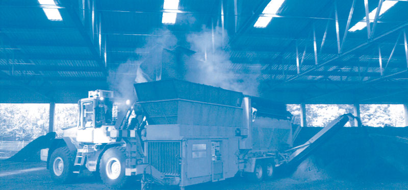 Ein großer Radlader in einer Lagerhalle schüttet organisches Material in eine Maschine, die Rauch oder Dampf freisetzt. Die Halle ist von hohen Decken und einer strukturierten Metallkonstruktion geprägt. Im Hintergrund sind Tageslichtöffnungen zu erkennen. Der Kontext könnte auf Arbeitsschutz und Prävention in der industriellen Verarbeitung hinweisen.