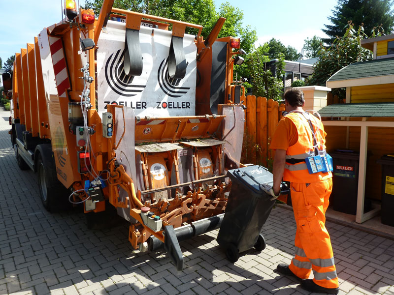 Ein Müllwerker in orangener Schutzkleidung leert eine schwarze Tonne in ein orangefarbenes Müllfahrzeug. Das Fahrzeug steht auf einer gepflasterten Straße in einem Wohngebiet, umgeben von Bäumen und Zäunen. Die Rückseite des Fahrzeugs zeigt den Schriftzug 'ZOELLER'. Dieses Bild verdeutlicht die Wichtigkeit von Sicherheits- und Gesundheitsschutzmaßnahmen im Bereich der Abfallentsorgung.