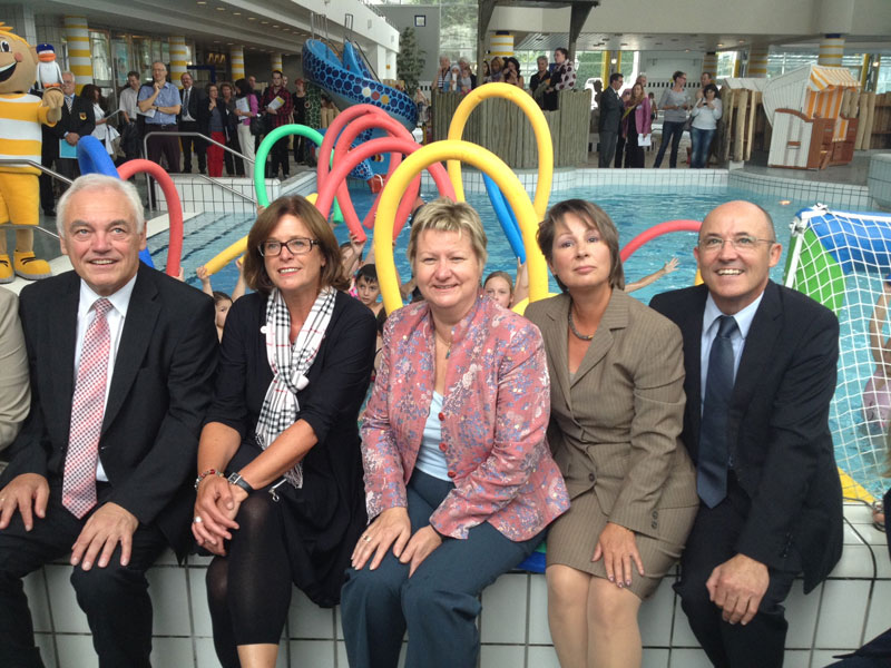 Fünf Personen in formeller Kleidung sitzen am Rand eines Schwimmbeckens. Im Hintergrund sind bunte Schwimmnudeln und spielende Kinder im Wasser zu sehen. Hinter den Kindern sind Zuschauer und Dekorationselemente, darunter ein Maskottchen, sichtbar. Die Veranstaltung scheint im Kontext von Arbeitsschutz und Prävention der Unfallkasse NRW zu stehen, was durch die Präsenz von Erwachsenen in Anzügen und die Einbindung eines Schwimmbades als Symbol für Sicherheit und Prävention verdeutlicht wird.