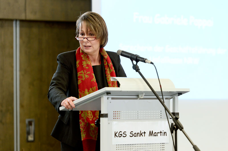 Eine Frau mit kurzem Haar und rotem Schal steht an einem Rednerpult und spricht ins Mikrofon. Auf dem Pult steht 'KGS Sankt Martin'. Im Hintergrund ist unscharfer Text zu erkennen. Die Szene findet in einem öffentlichen Raum statt. Der Kontext ist eine Veranstaltung, die im Zusammenhang mit Arbeitsschutz und Prävention der Unfallkasse NRW steht.