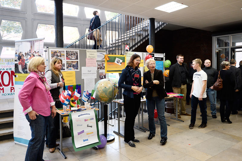 Ein Messebereich in einem offenen Raum mit Besuchern. Mehrere Stände sind mit Informationsmaterialien, Broschüren und Dekorationen ausgestattet. Ein Tisch zeigt bunte Flaggen kleiner Länder auf dem Tisch, daneben befindet sich ein Globus. Auf Plakaten an der Wand sind Texte und Bilder zu sehen. Menschen stehen, schauen sich um und unterhalten sich. Der Raum hat große Fenster im Hintergrund, die natürliches Licht hereinlassen. Kontinuierliche Themen wie Arbeitsschutz und Prävention werden thematisiert.