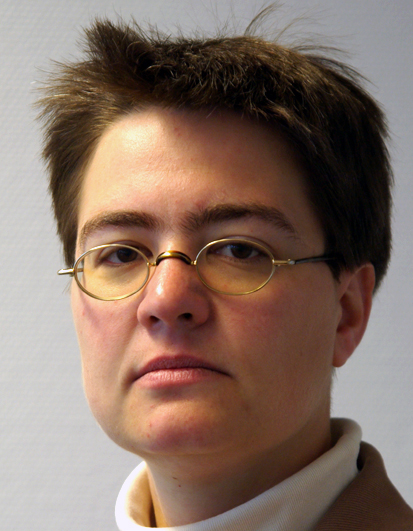 Nahaufnahme einer Person mit kurzen, dunklen Haaren und einem neutralen Gesichtsausdruck. Die Person trägt eine kleine, runde Brille mit goldfarbenem Rahmen und ein weißes Oberteil unter einem hellbraunen Blazer. Der Hintergrund ist schlicht und hell. Kontext für die Unfallkasse NRW könnte auf Arbeitsschutz und Prävention hinweisen, etwa durch die Darstellung einer Expertin oder eines Experten in diesem Bereich.