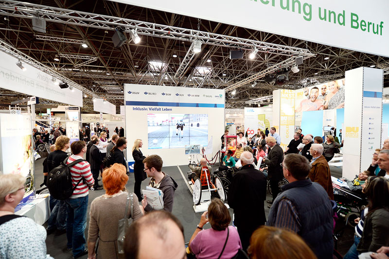 Eine große Messehalle ist gefüllt mit vielen Besuchern, die sich eine Präsentation zum Thema Arbeitsschutz und Inklusion ansehen. Im Hintergrund sind Stände verschiedener Organisationen zu sehen, darunter auch eine Informationstafel, die auf die Unfallkasse NRW hinweist. Eine große Leinwand zeigt eine visuelle Präsentation über Inklusion im Arbeitsleben. Die Atmosphäre ist geschäftig und informativ, mit einem Fokus auf Prävention und Diversität am Arbeitsplatz.