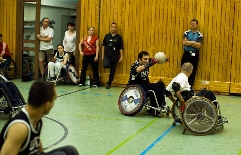 Mehrere Sportler in Rollstühlen spielen Rollstuhl-Rugby in einer Turnhalle. Zwei Spieler im Vordergrund sind im intensiven Wettkampf um den Ball. Zuschauer und Schiedsrichter stehen am Spielfeldrand. Das Bild steht im Kontext der Unfallkasse NRW, die sich für Arbeitsschutz und Prävention insbesondere auch im Rahmen von inklusiven Sportveranstaltungen einsetzt.