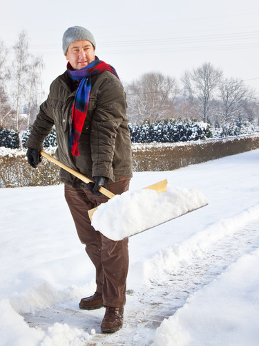 Eine Person in warmer Kleidung, einschließlich Mütze, Handschuhen und Schal, schaufelt Schnee von einem Gehweg. Der Hintergrund zeigt verschneite Bäume und Hecken. Dies unterstreicht die Bedeutung des Arbeitsschutzes beim Schneeräumen, um Unfälle zu vermeiden. Kontextualisiert für die Unfallkasse NRW im Bereich Arbeitsschutz und Prävention.